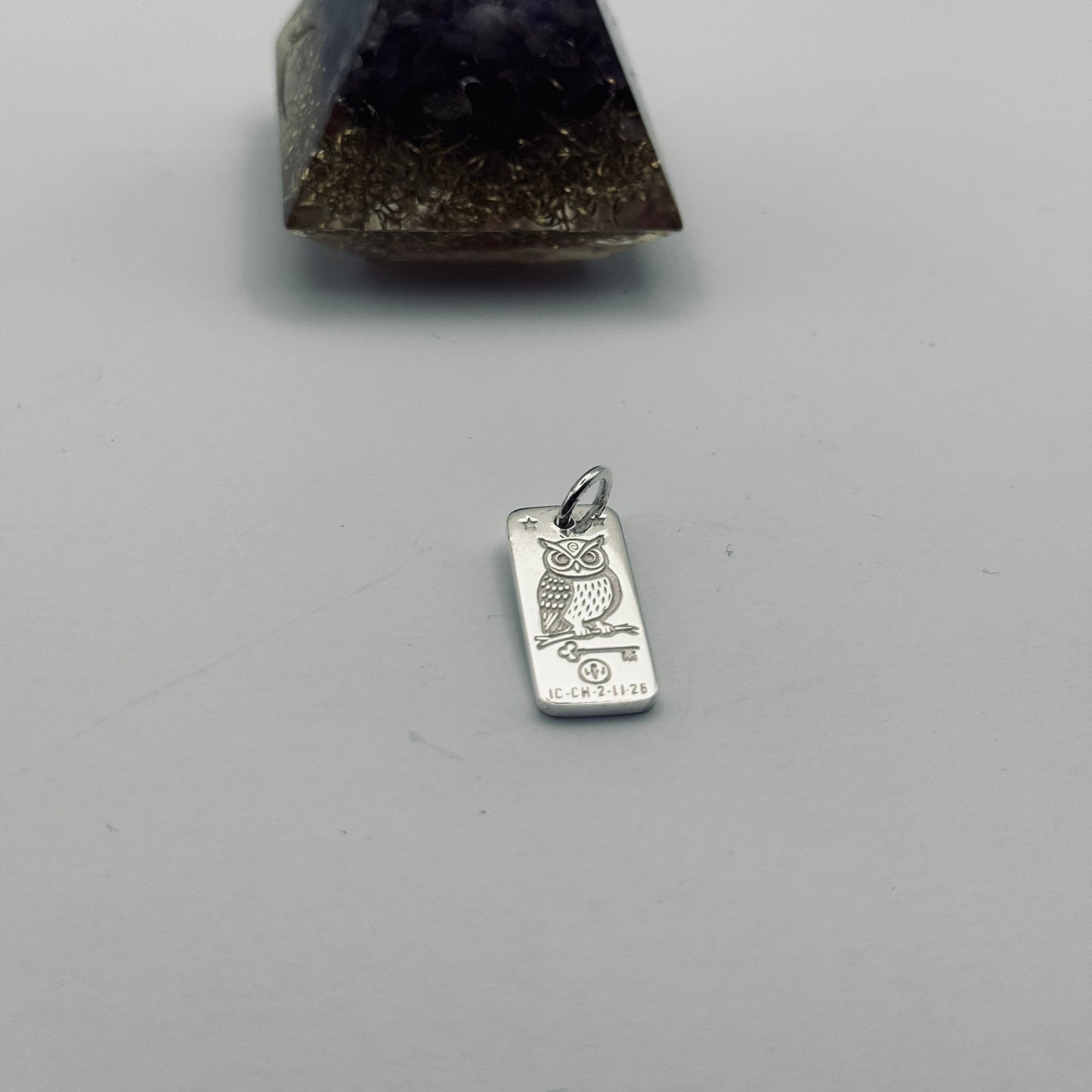 A silver owl pendant charm engraved with 'ECO FASHION' and a small silver ring at the top, shown below a dark purple amethyst and gold glitter pyramid crystal on a light gray background.